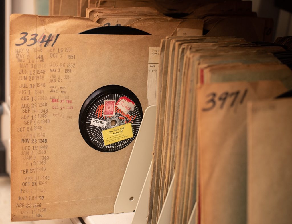 Close up view of a series numbered 78 rpm record discs in a paper sleeves on a metal shelf with dividers that help the discs to stand vertically. A disc, numbered "3341" in bold black ink, is pulled out slightly from the rest of the items on the shelf to show that its sleeve has been stamped several times with various dates ranging from May, 2, 1948 to December 27, 1964.