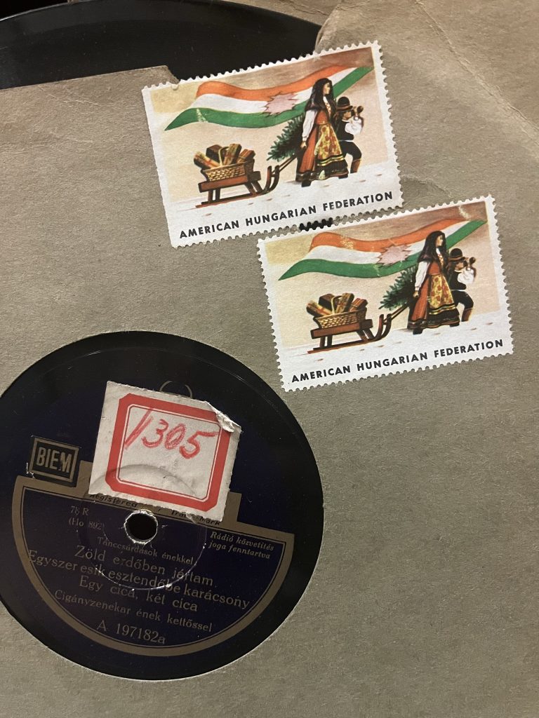 Close up of 78 rpm record disc in a paper sleeve that has its center cut in a circle to show the record's title label, which is printed in Hungarian. A sticker with an identification number on it partially covers the top part of disc's label. Two "American Hungarian Federation" Christmas sticker-like postage stamps are stuck to the paper sleeve above and to the right of the circular opening that shows the disc's label.
